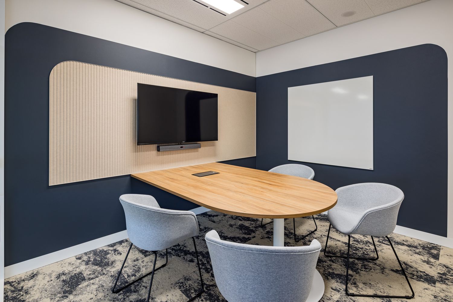 Small modern meeting room with an oval wooden table, light gray chairs, wall-mounted TV, whiteboard and navy accent walls.
