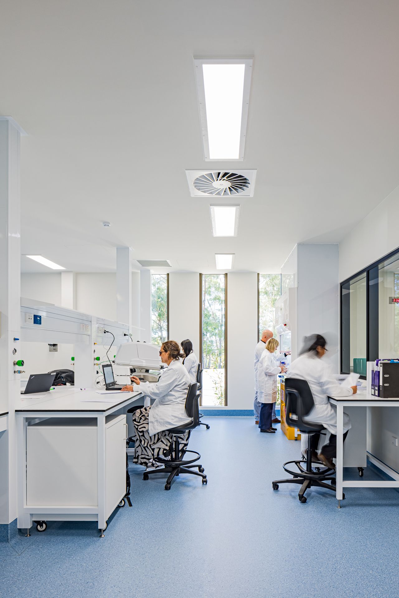 Bright modern laboratory with scientists in white coats working at benches under skylights, with large windows overlooking trees.