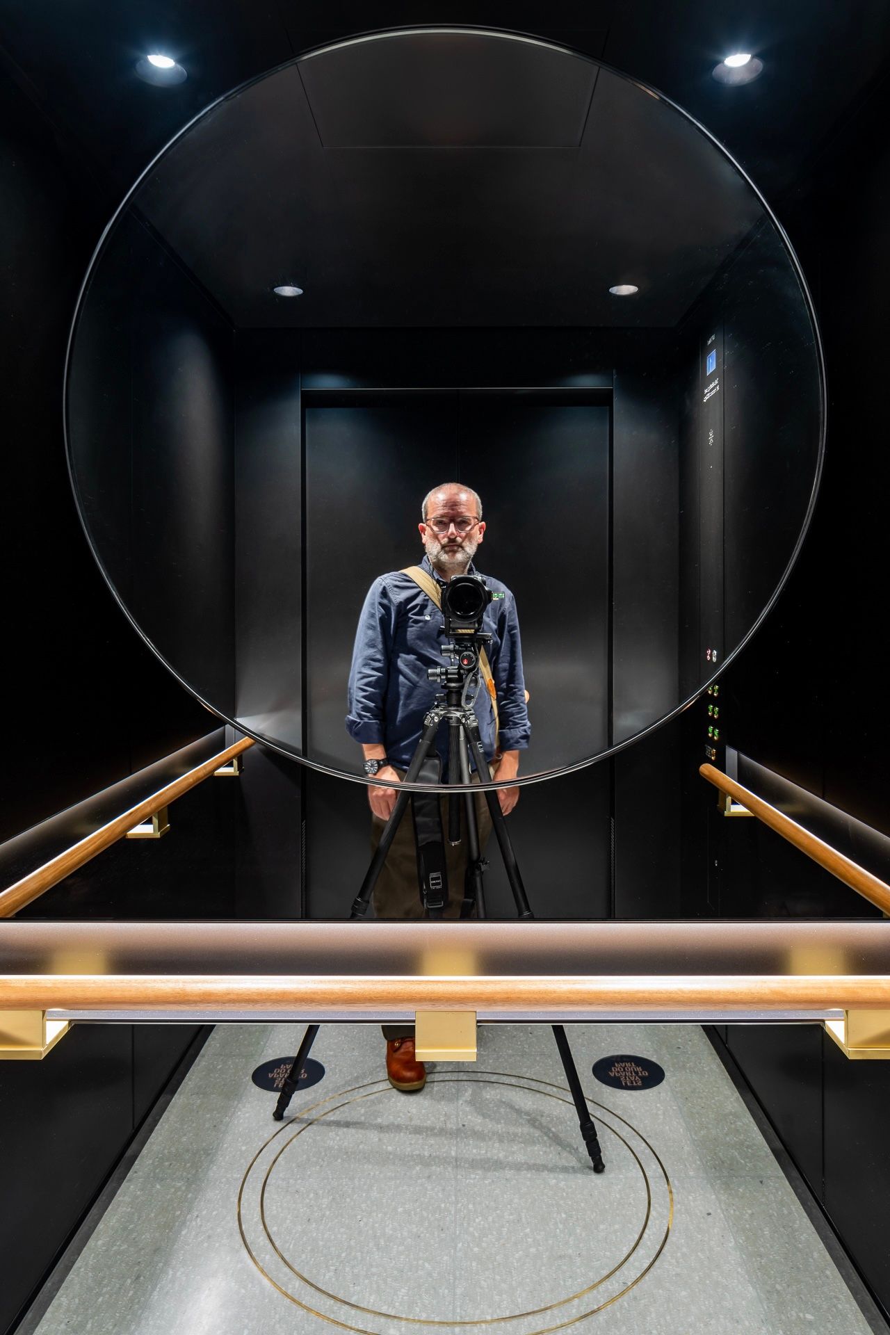 Photographer with a tripod-mounted camera reflected in a round mirror inside a black elevator with brass handrails.