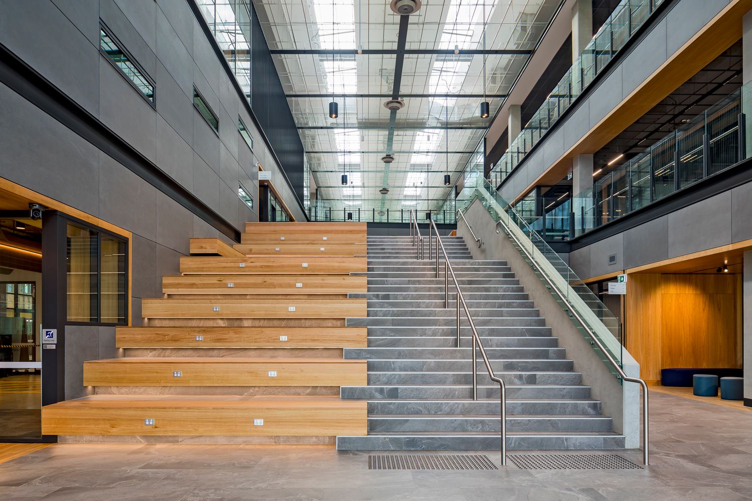 Wide concrete staircase beside timber tiered seating in a modern, light-filled atrium with glass balustrades and upper-level walkways.