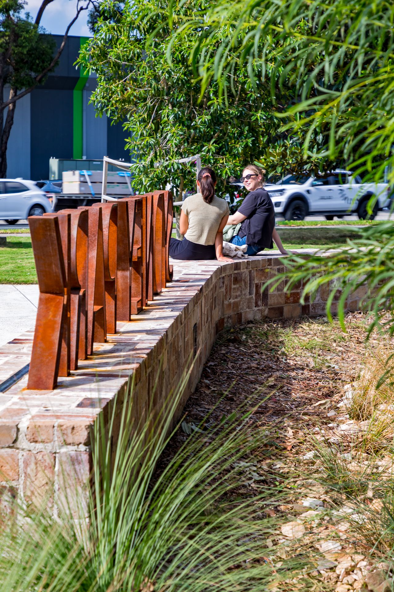 Two people sit on a curved sandstone wall beside rusty steel letters, with native grasses and parked cars beyond.