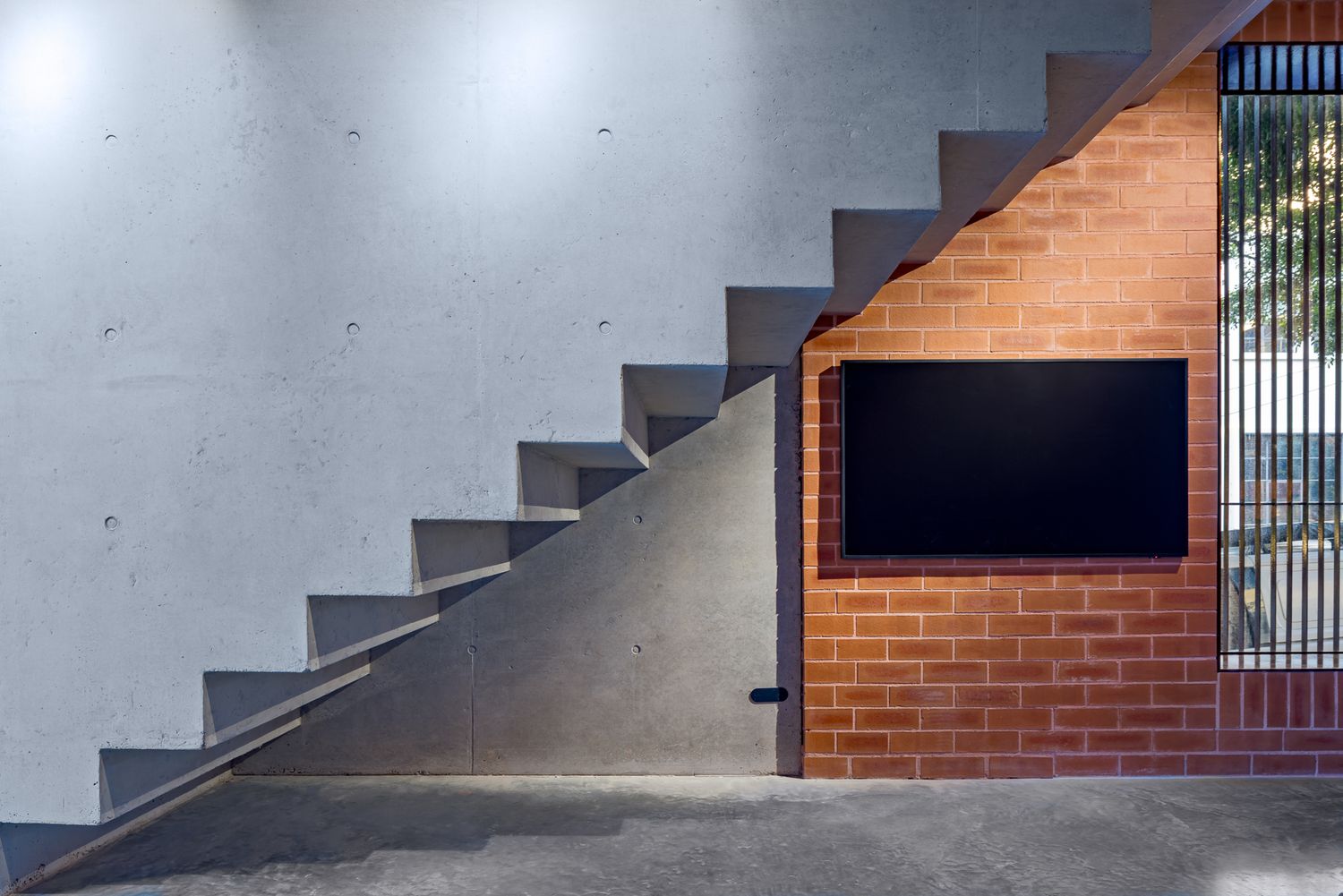 Raw concrete staircase beside a red brick wall with a wall-mounted flat screen TV in a minimalist architectural interior.