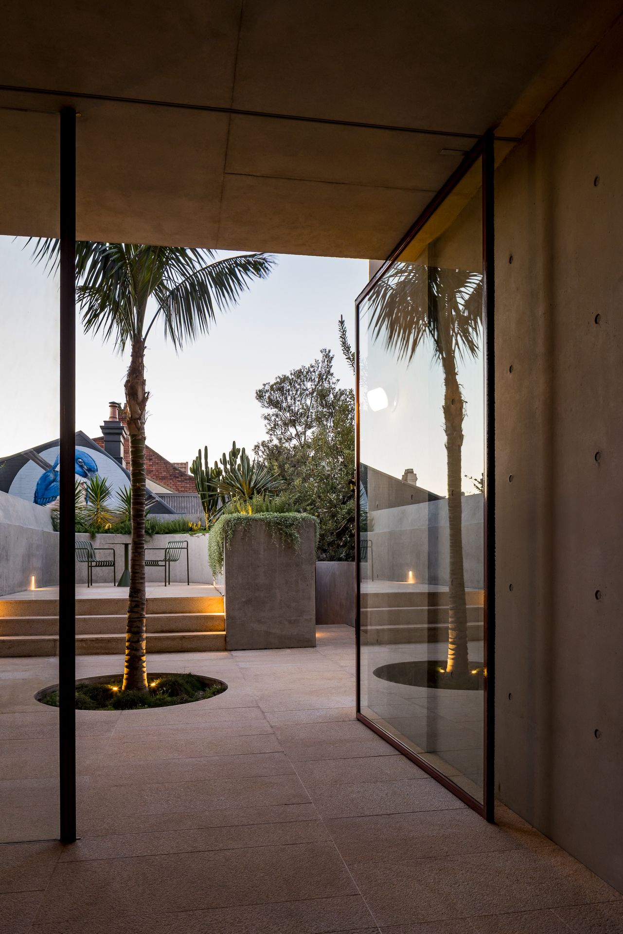 Glass pivot door opening from a concrete interior onto a paved courtyard with a palm tree and softly lit steps at twilight.