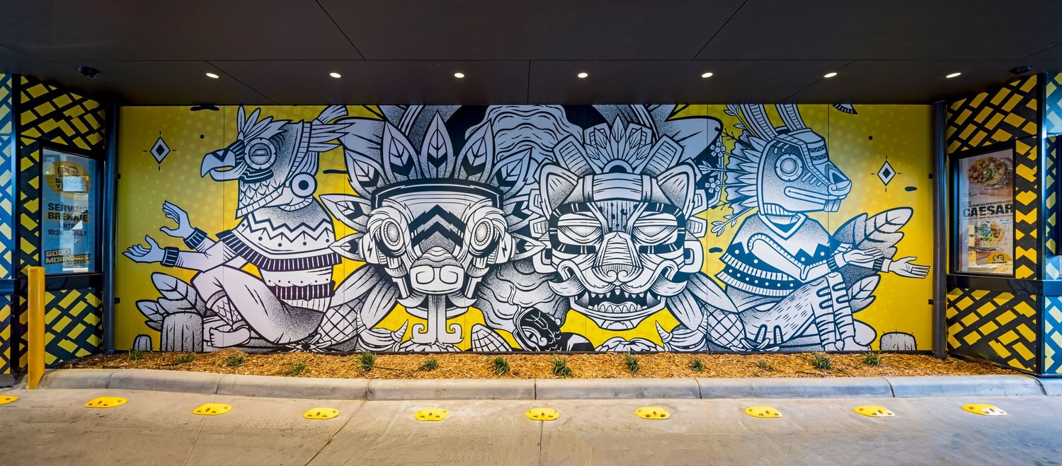 Colorful street art mural of stylized masked animal faces in blue, gray and yellow spans an exterior wall beside a concrete walkway.