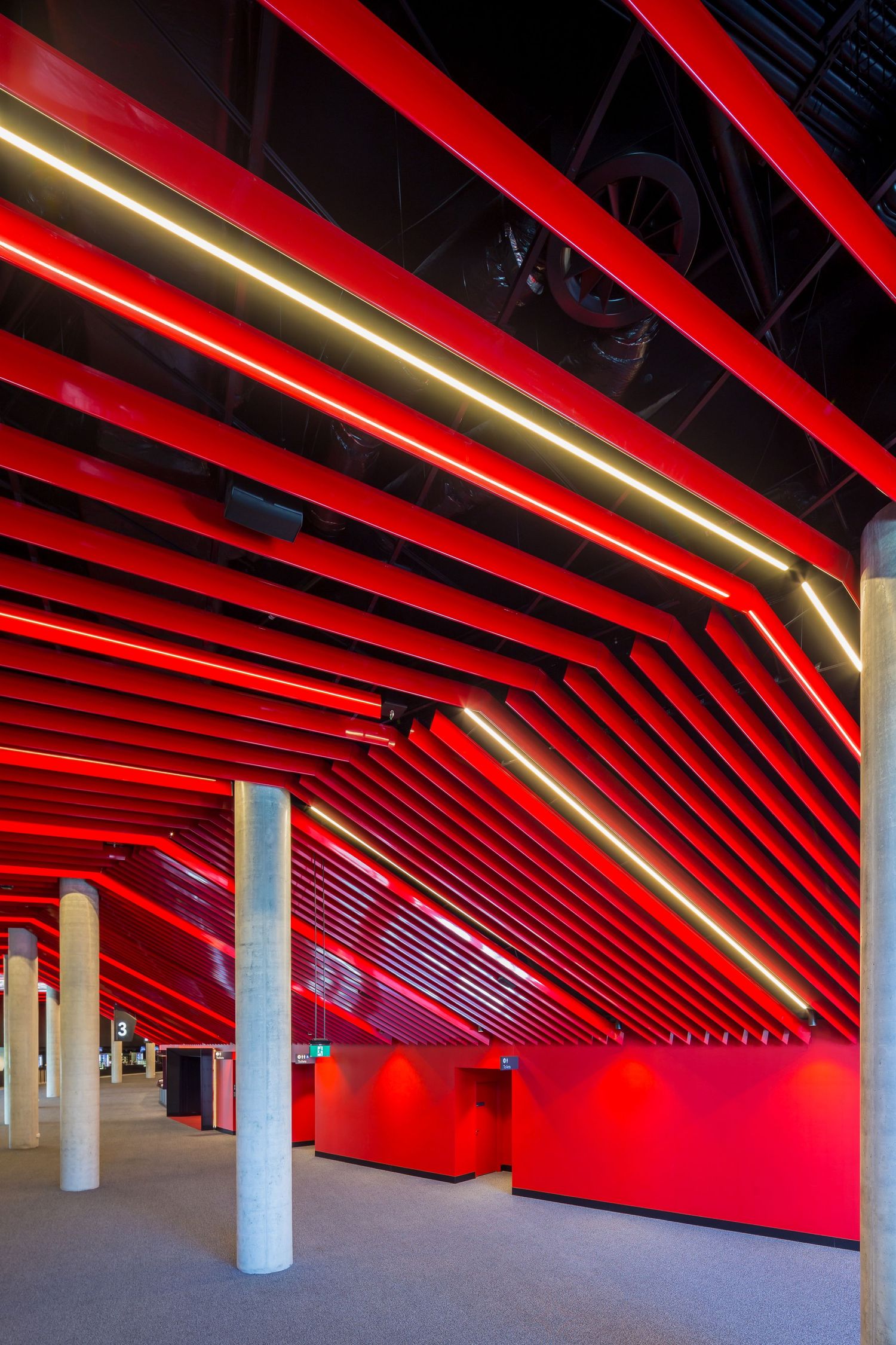 Close view of a vivid red angled ceiling with warm linear lights running above concrete columns in a modern event space.