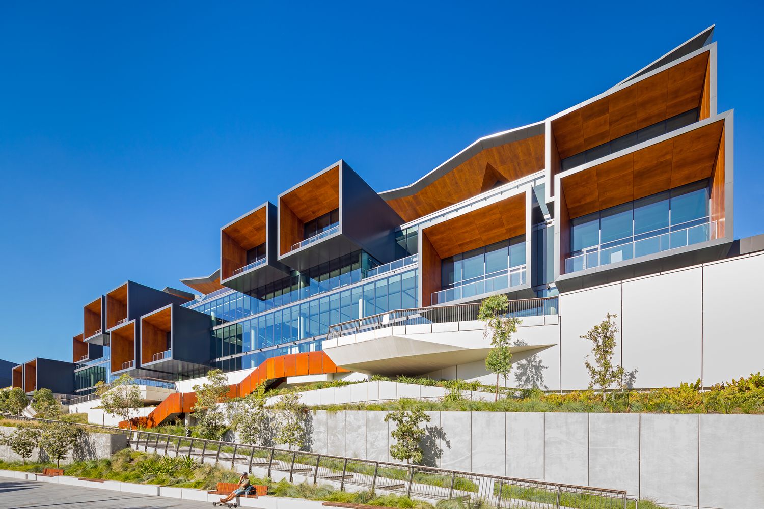 Stepped modern apartment-style building with timber-lined balconies, glass walls and landscaped gardens under a clear blue sky.