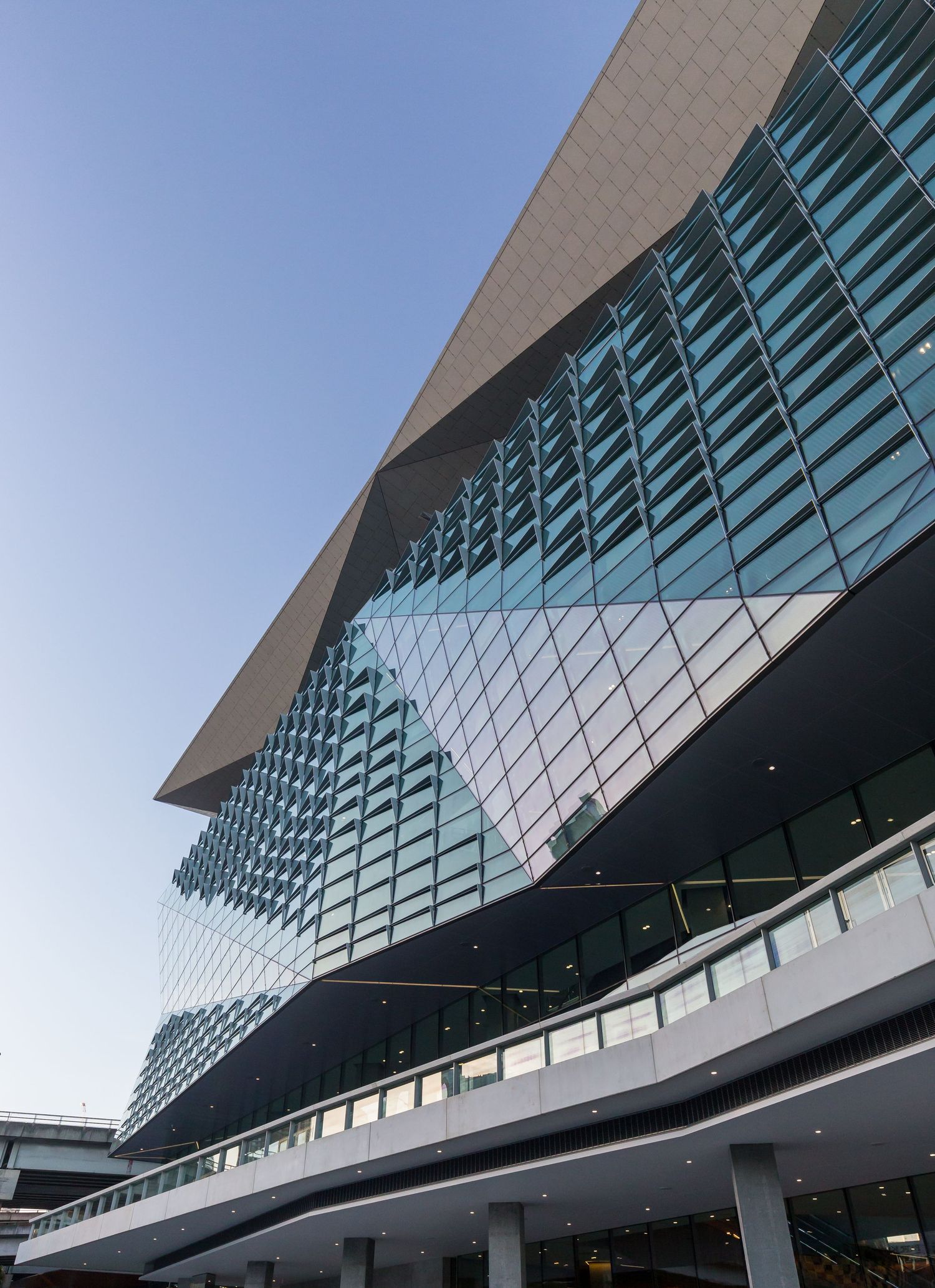 Upward view of a contemporary glass facade with angular geometric panels and a sloping metallic roof against a clear sky.