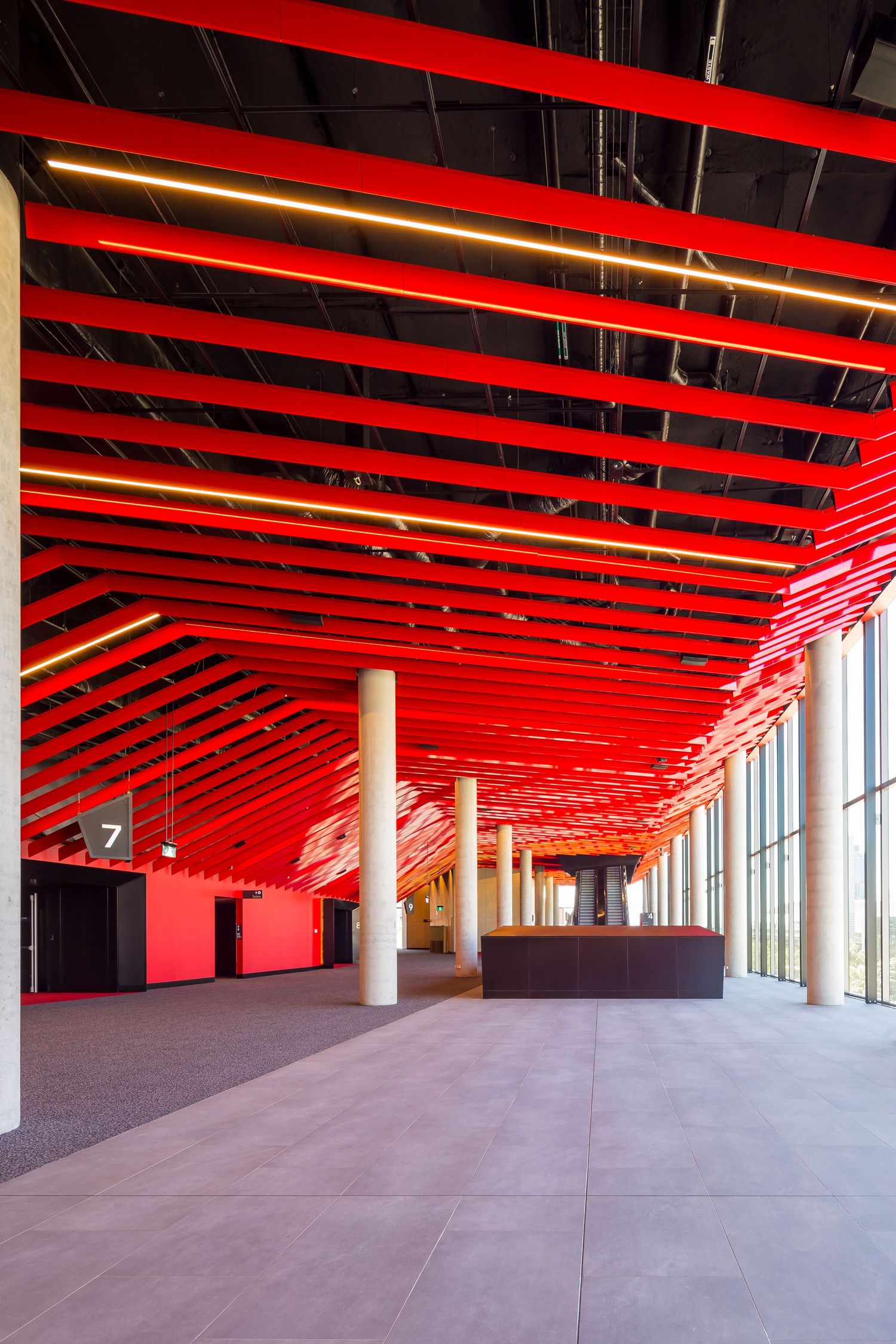 Spacious event foyer with a bold red slatted ceiling, linear lighting, concrete columns and glass walls overlooking the city.