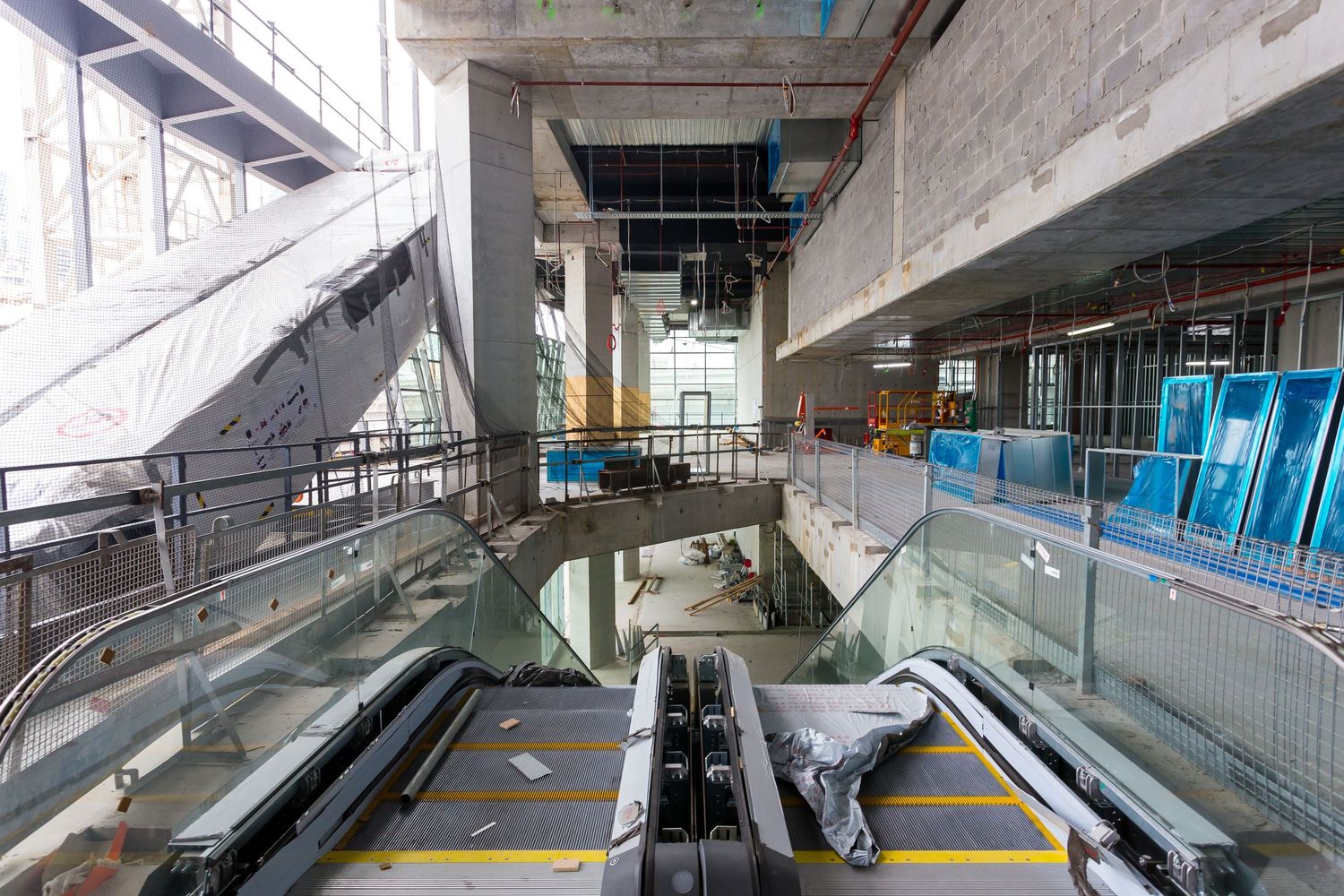 Unfinished building interior with glass-balustrade escalators, concrete columns, and protective wrapping during construction.