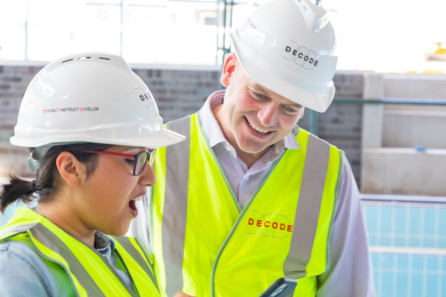 Two smiling construction workers in white hard hats and yellow high-visibility vests review plans together at a building site.