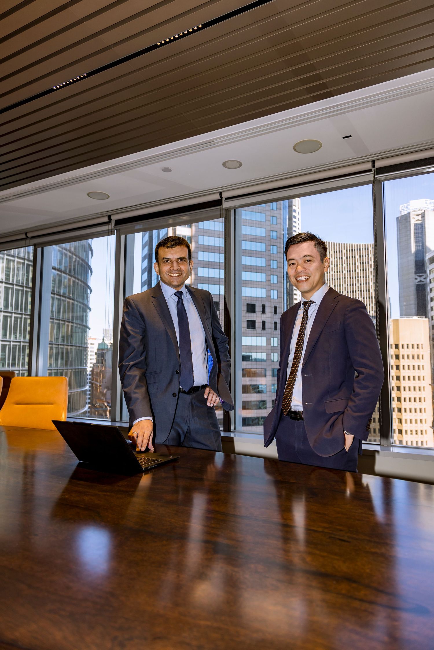 Two smiling businessmen in suits stand beside a laptop on a polished boardroom table with city skyscraper views through large windows.