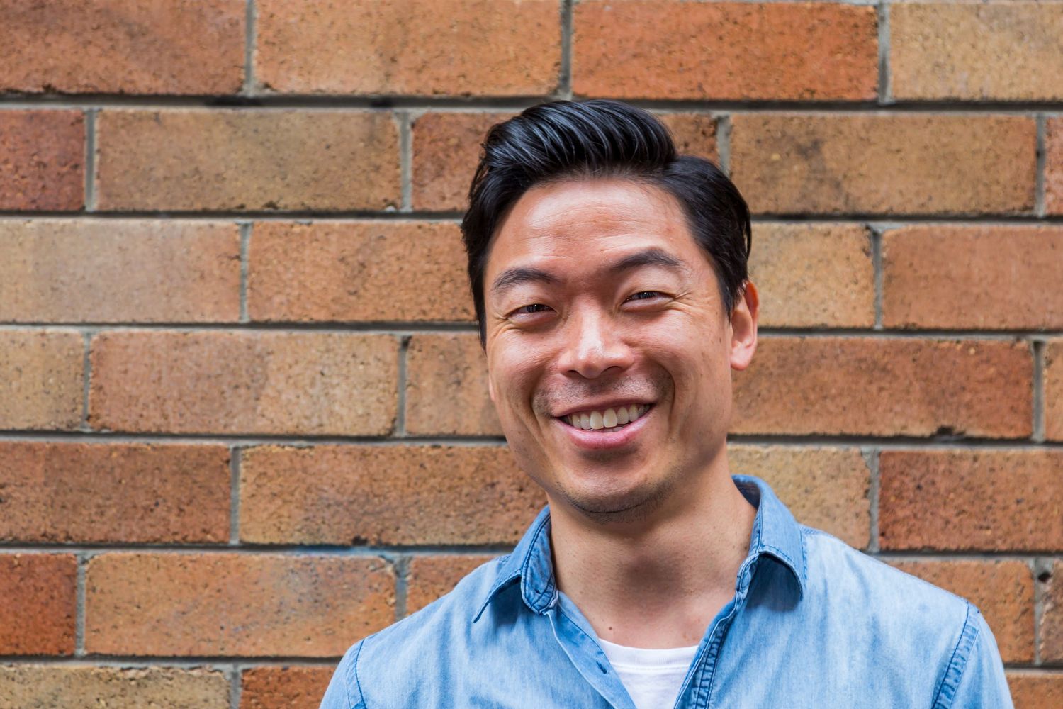 A smiling man with dark hair wearing a denim shirt over a white tee stands in front of an orange brick wall.