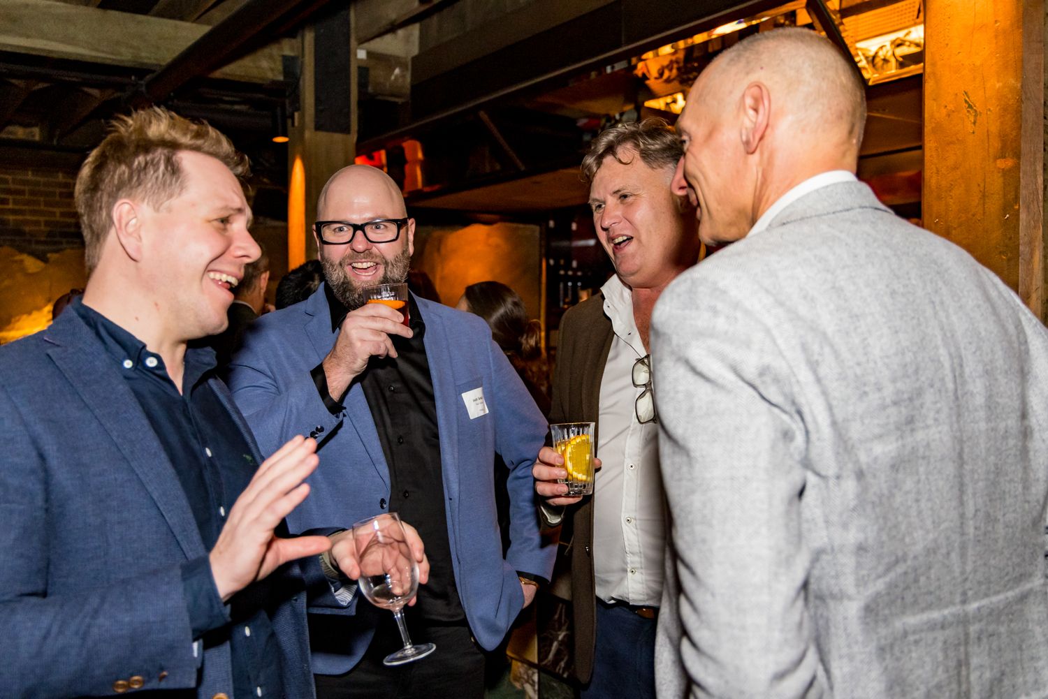 Four men in smart casual clothing chat and hold drinks at a social networking event in a warmly lit venue.