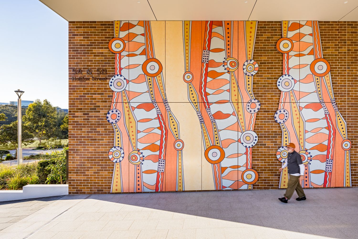 Pedestrian walks past a vibrant Indigenous art mural with orange and blue circles on a brick facade.