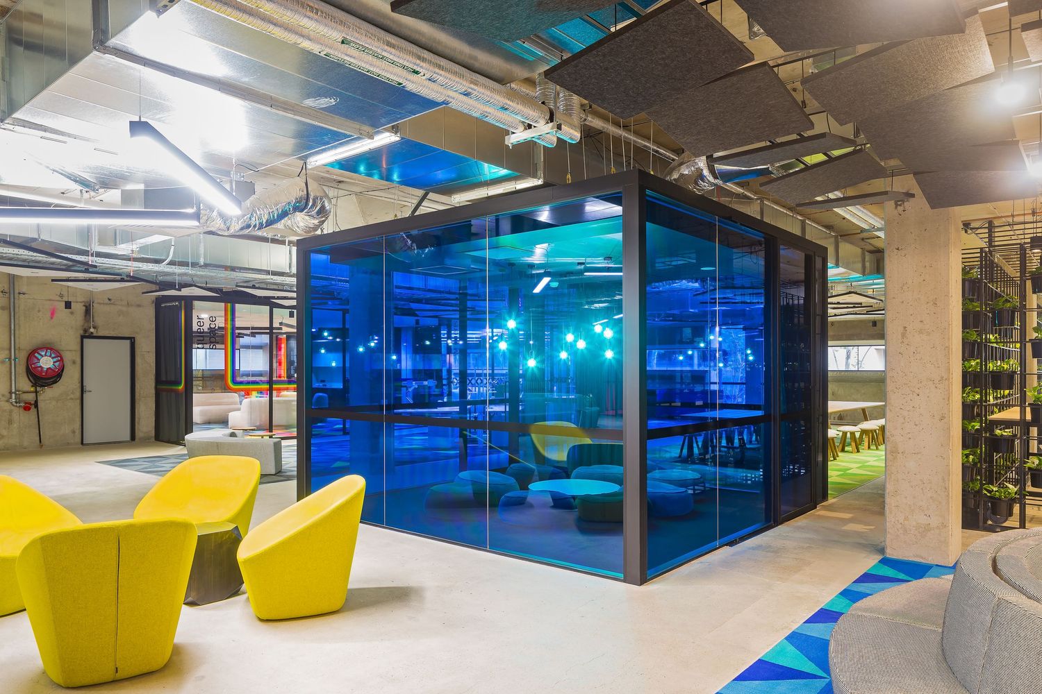 Contemporary office space with a blue glass meeting pod, bright yellow lounge chairs and exposed industrial ceiling ducts.