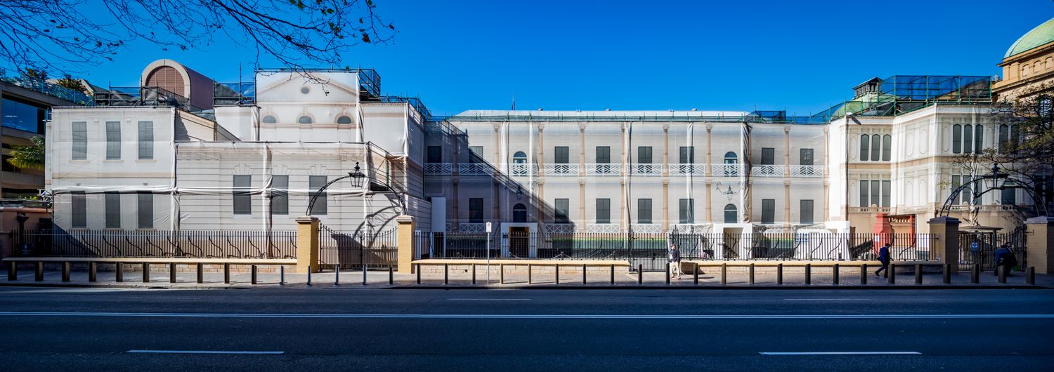 Wide panoramic view of a restored heritage building facade with symmetrical windows beside a street under a clear blue sky.
