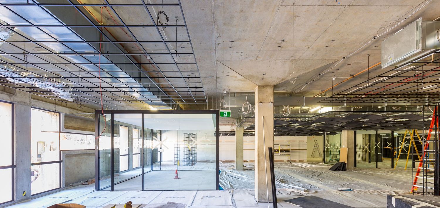Interior of a commercial building under construction with exposed concrete ceiling, glass partitions and a ladder on the site floor.