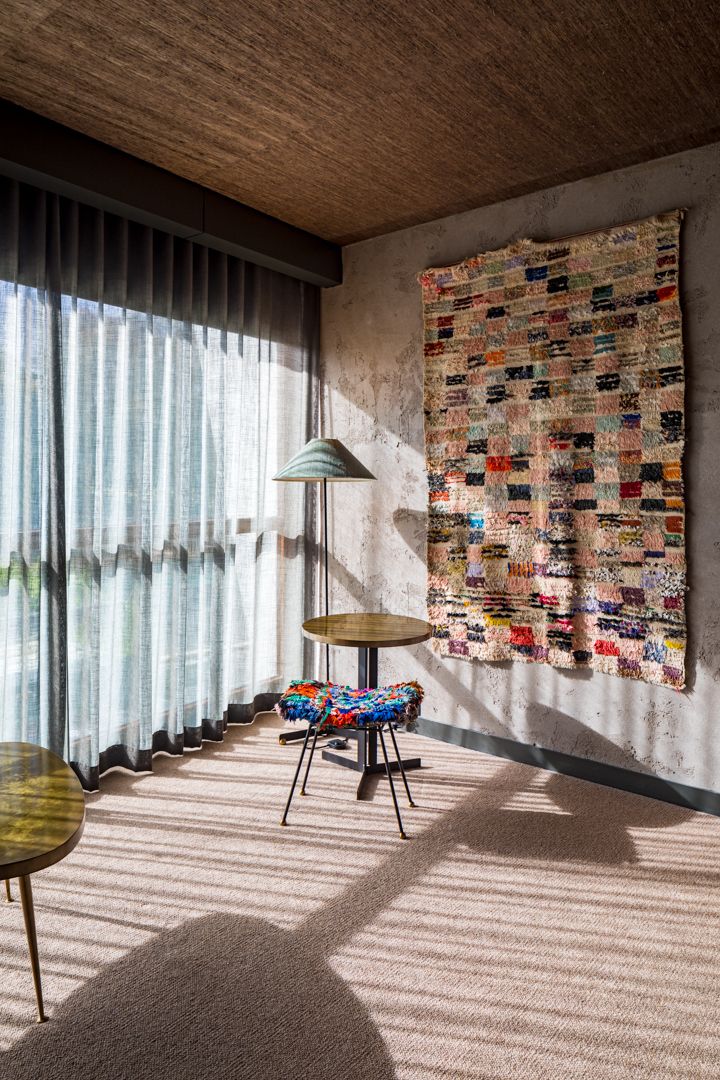 Cozy interior corner with a colorful woven rug on a concrete wall, a patterned stool, side table, floor lamp and sheer curtains.