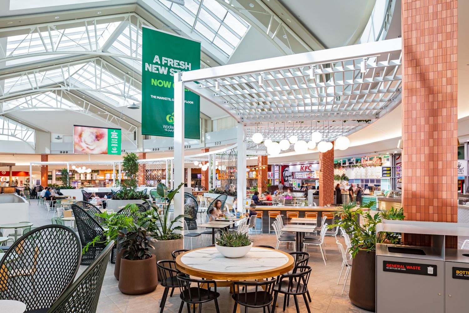 Bright shopping centre food court with skylights, potted plants, wooden tables, black chairs and a green fresh new menu banner.