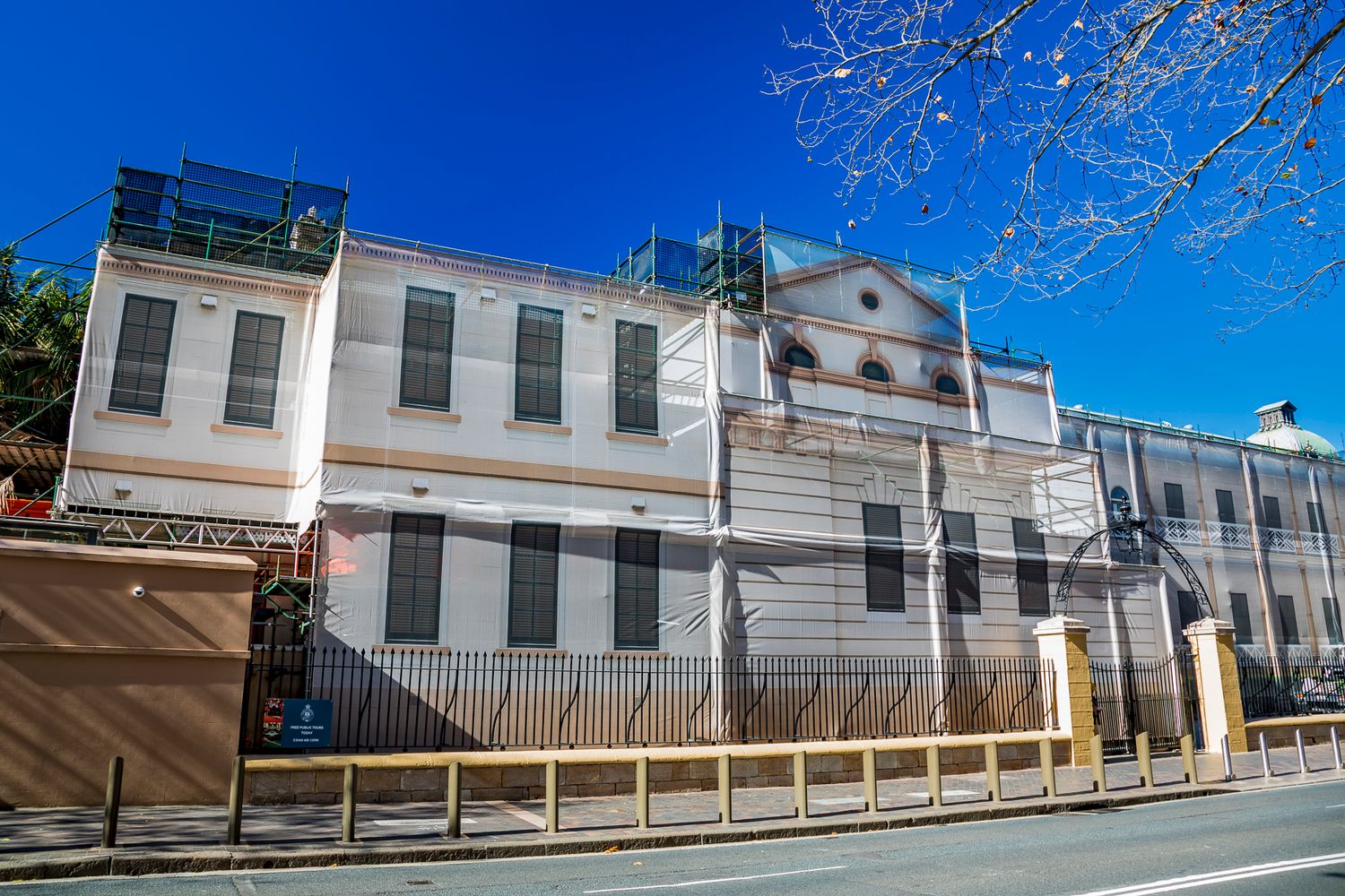 Historic cream-colored building with scaffolding and an iron fence along a sunny street under a blue sky.
