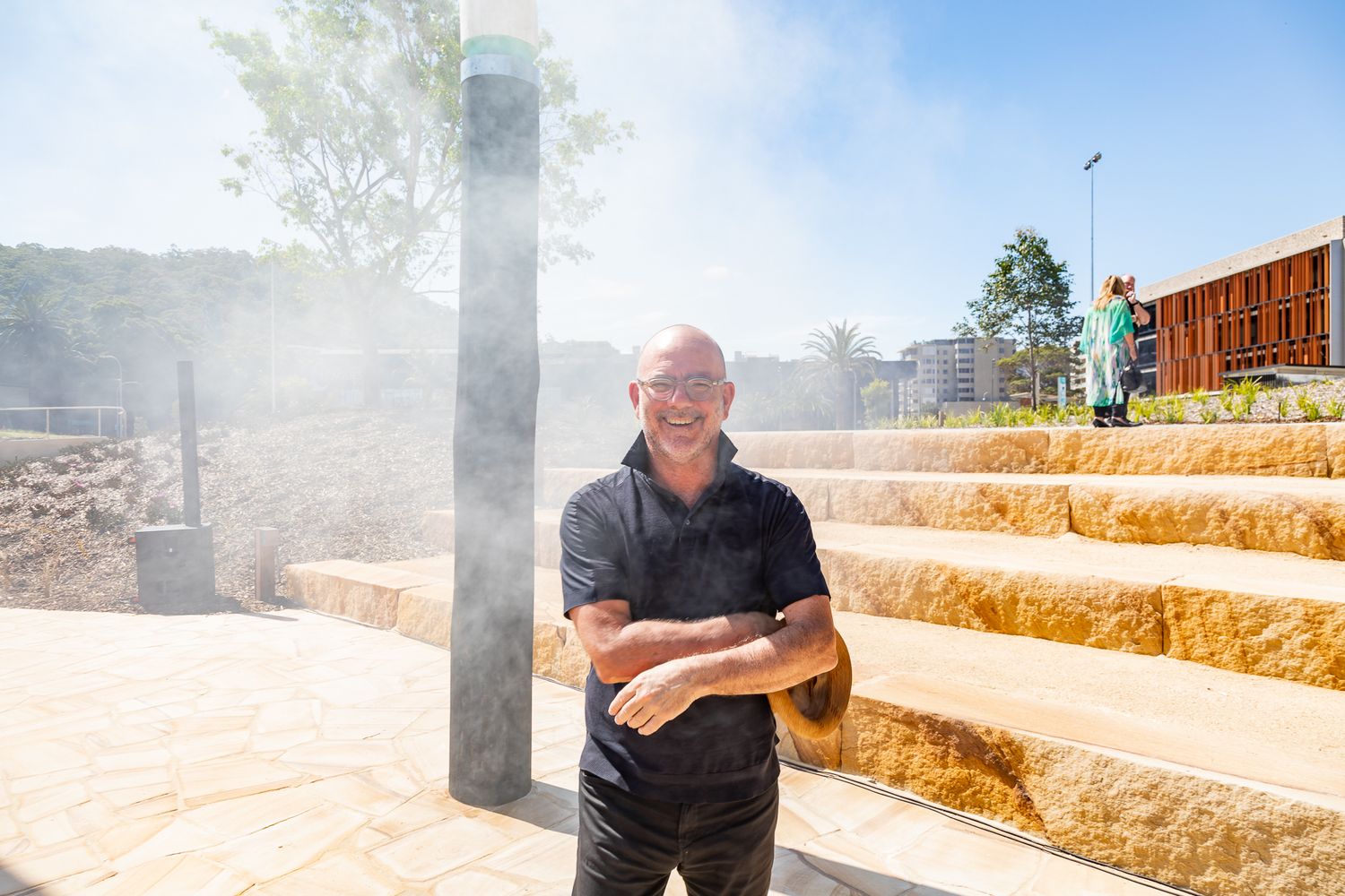 Smiling bald man in a black shirt stands with folded arms on sandstone terraced steps as mist drifts through a sunny public park.