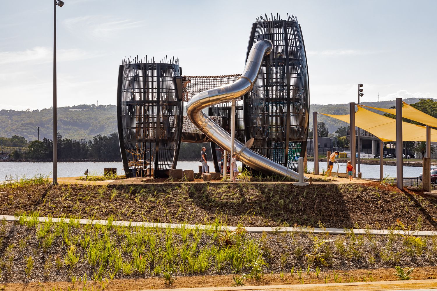 Large curved metal slide connects two lattice play towers at a lakeside playground, with shade sails and rolling hills beyond.