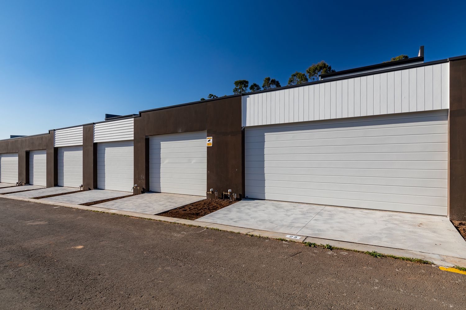 Row of townhouse garages with white roller doors, brown brick walls and concrete driveways beside a paved lane.