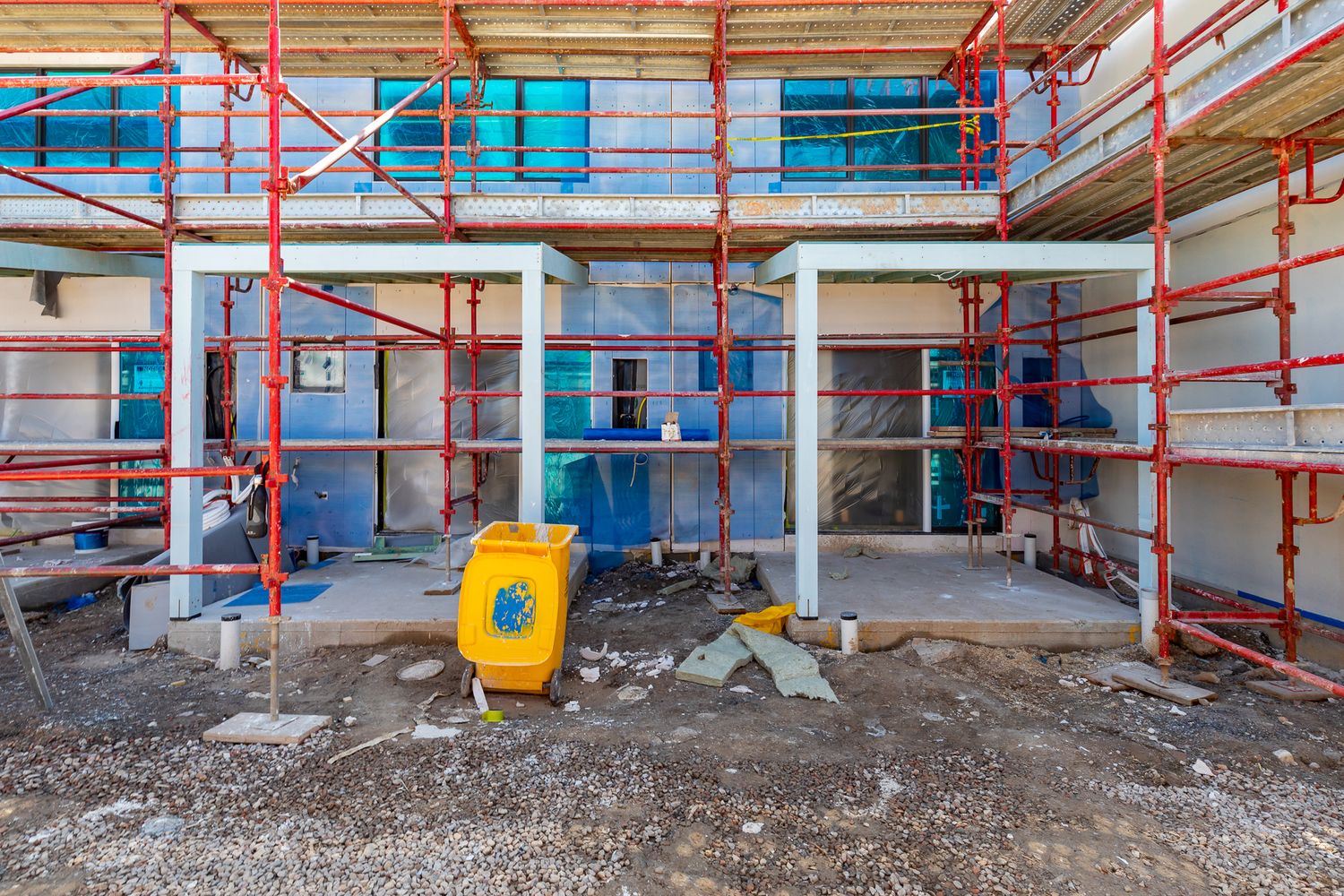 Red scaffolding around an unfinished townhouse block with a yellow bin and construction debris scattered across the ground below.