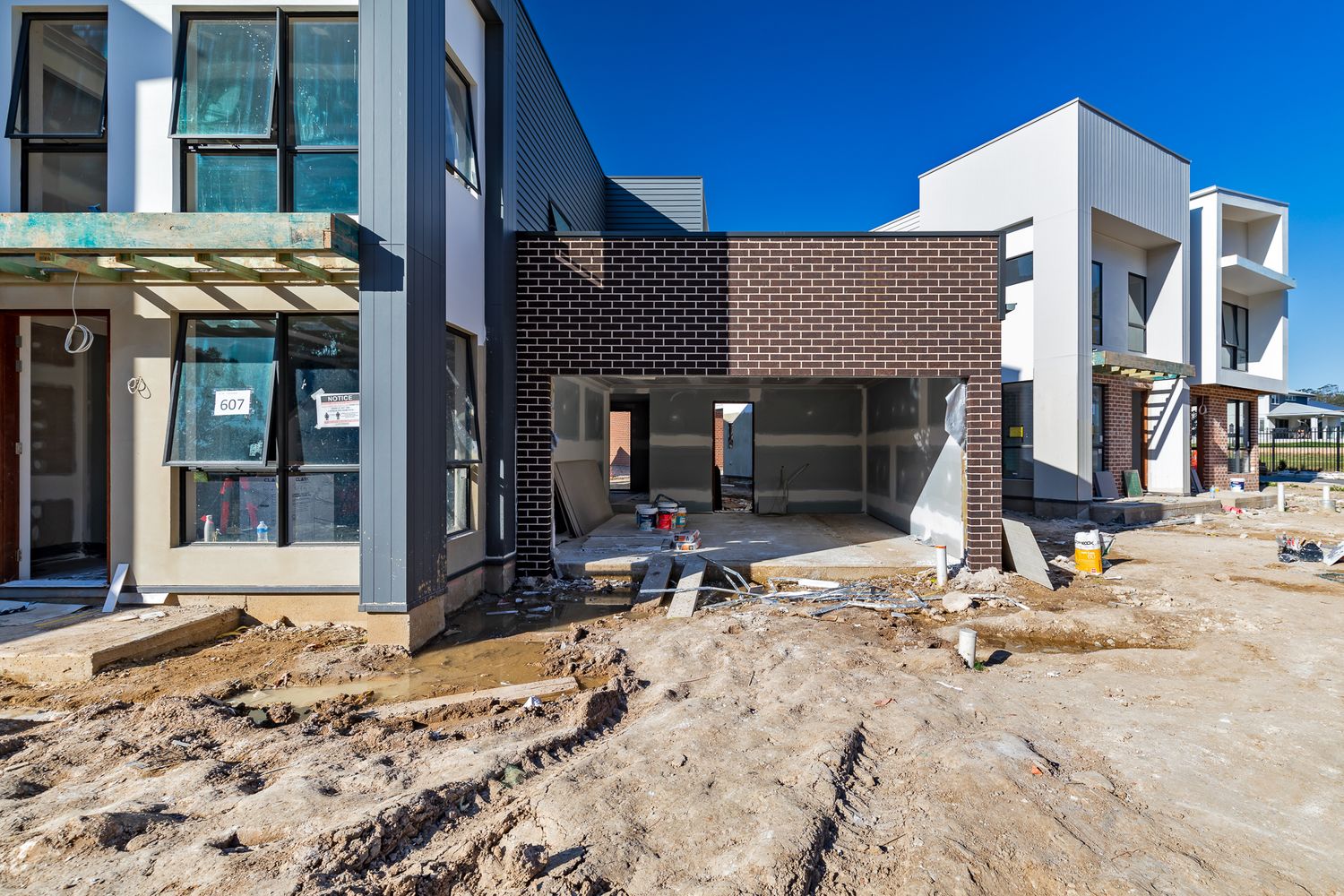 Modern townhouse under construction with brick and grey cladding, an open entry and muddy ground in the foreground.