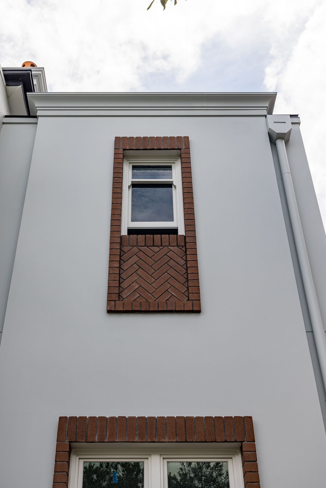 Grey rendered house facade with a tall narrow window framed by herringbone brick detailing and a downpipe.