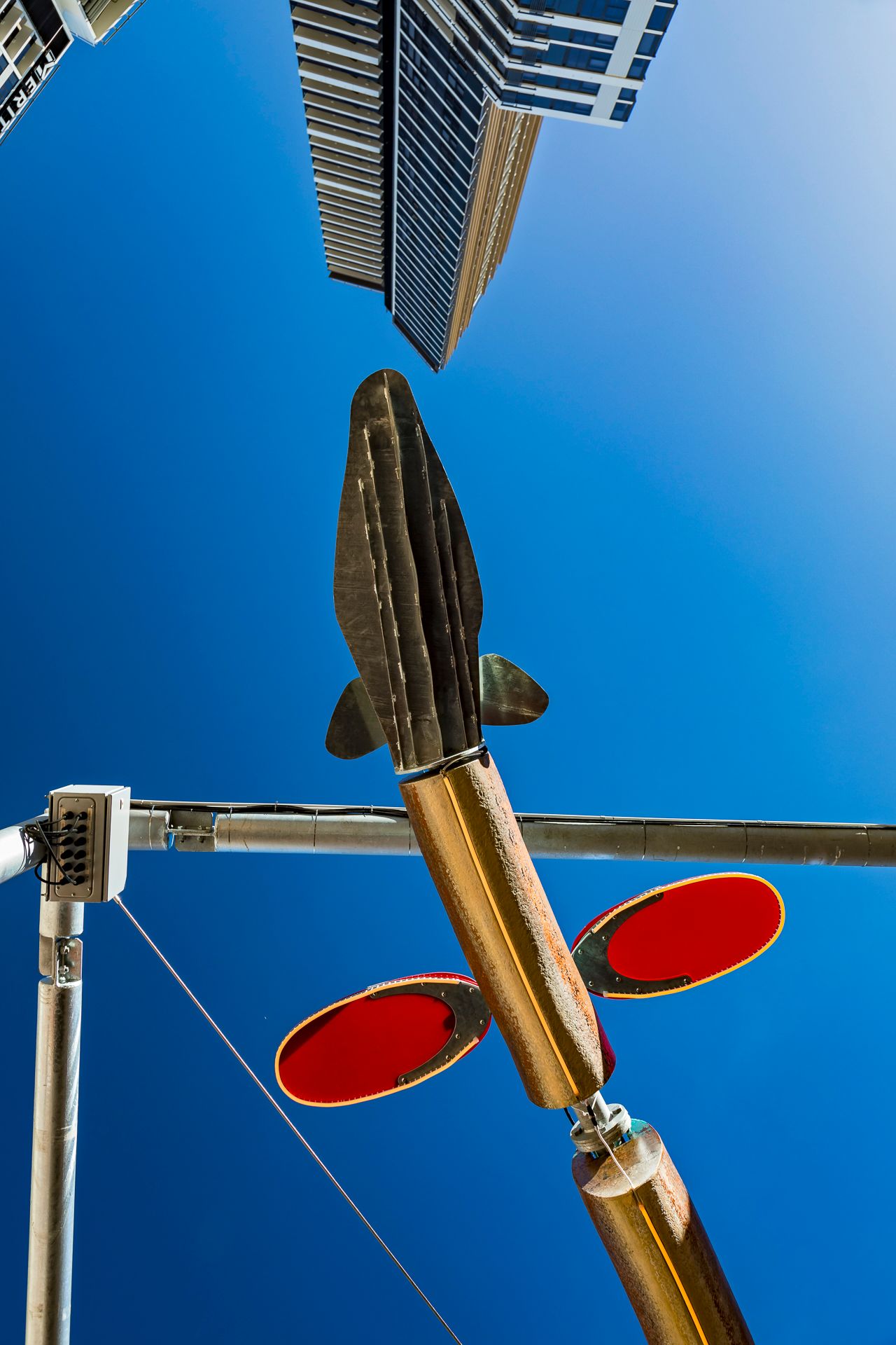 Low-angle view of a fish-shaped bronze sculpture with red oval discs suspended from steel poles between skyscrapers and blue sky.