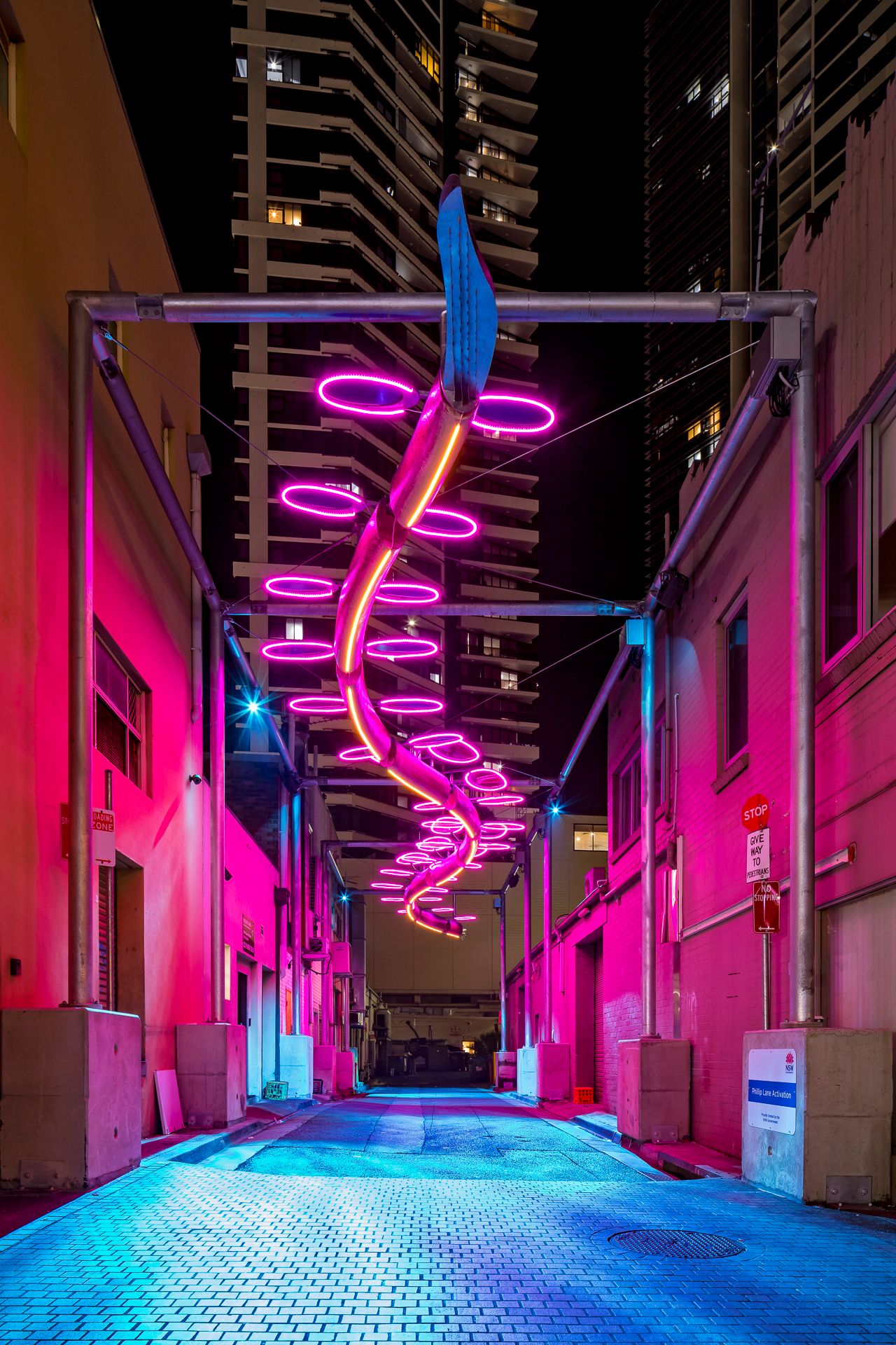 Pink and orange neon rings coil above a bright magenta alley, creating a vibrant nighttime urban art scene.