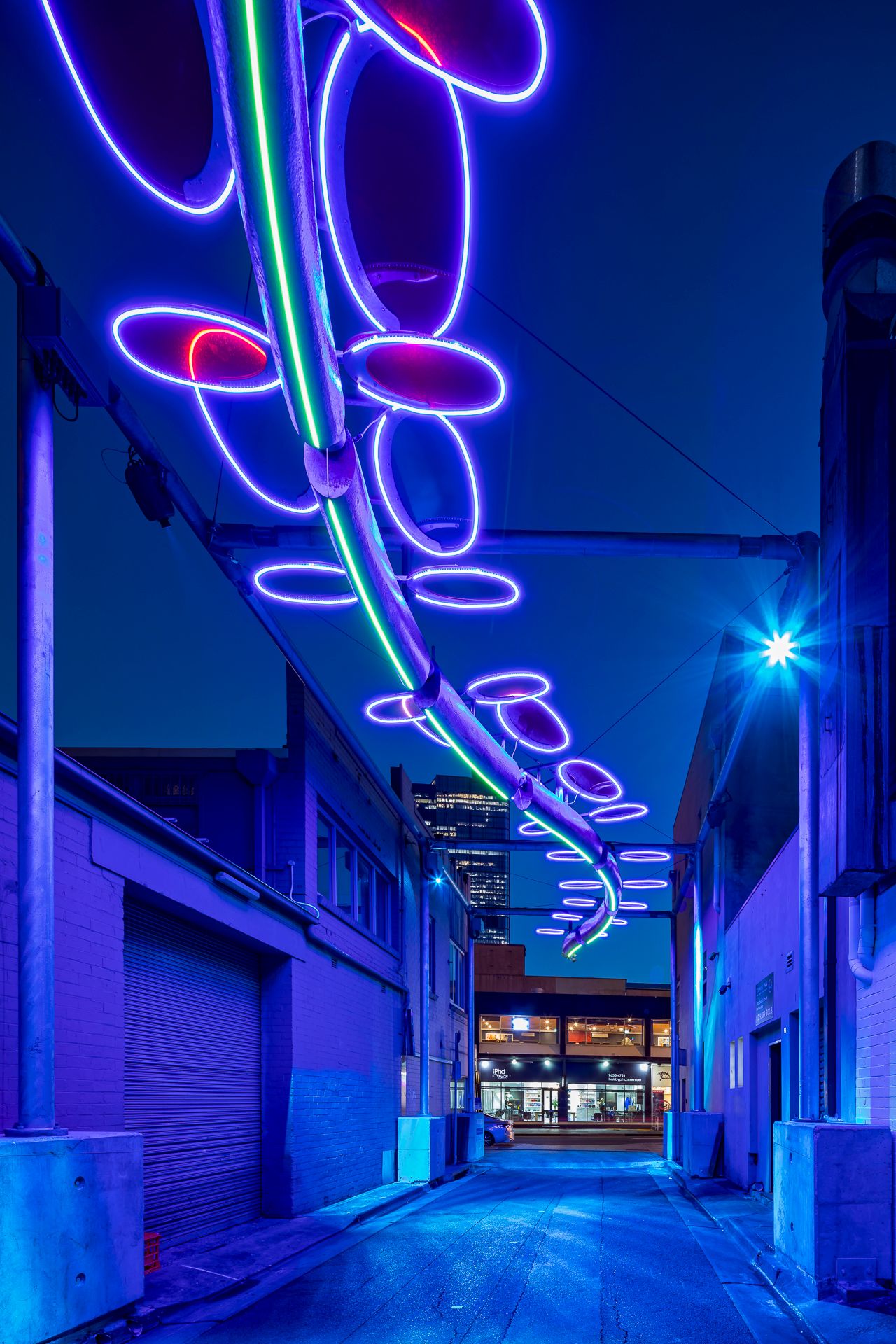 Blue and purple neon oval light sculpture with green accents hanging over a dark city alleyway at night, skyscraper in the background.