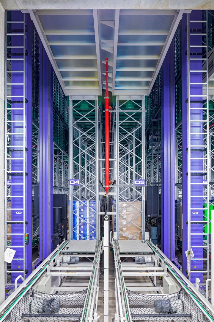 Automated high-bay warehouse aisle with tall blue steel racking, a red-lined shuttle mast, and conveyor tracks in the foreground.