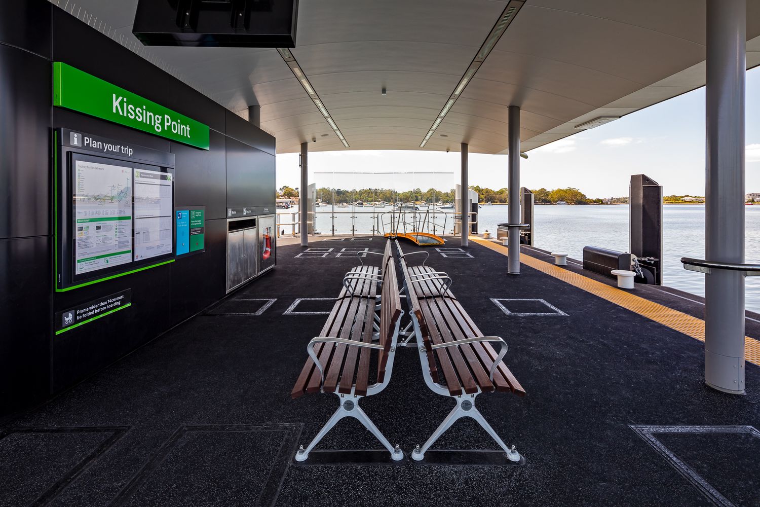 Rows of wooden benches under the covered Kissing Point wharf waiting area, with a route map display and river views beyond.
