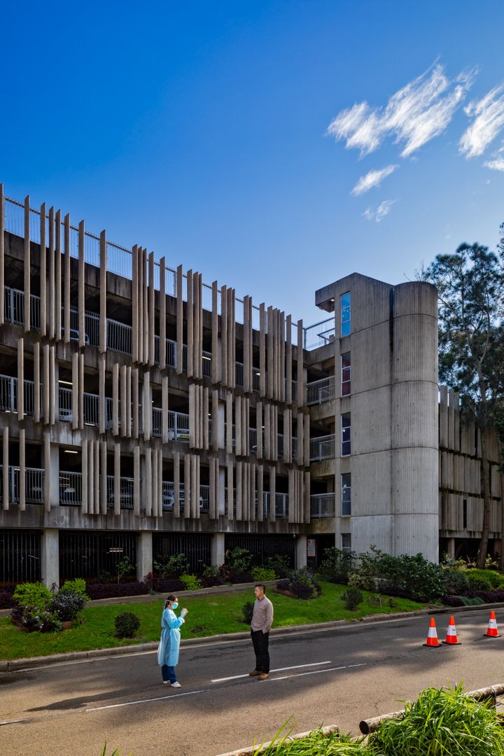 Brutalist concrete car park with vertical timber-style screens and landscaped garden, with two people in protective gear on the road.
