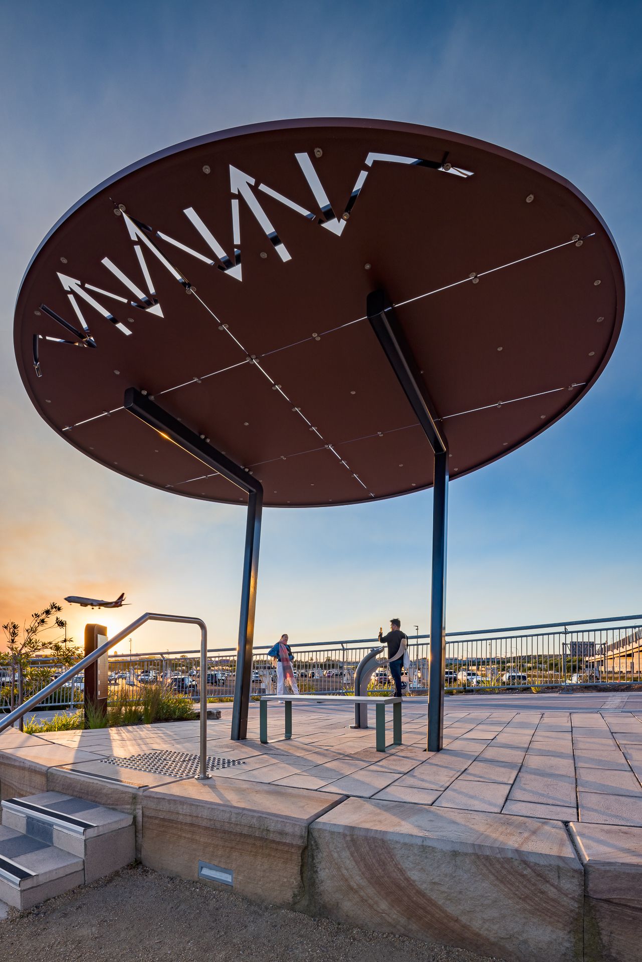 Large circular canopy with cut-out lettering shelters people on a paved lookout as an airplane flies across the sunset sky.