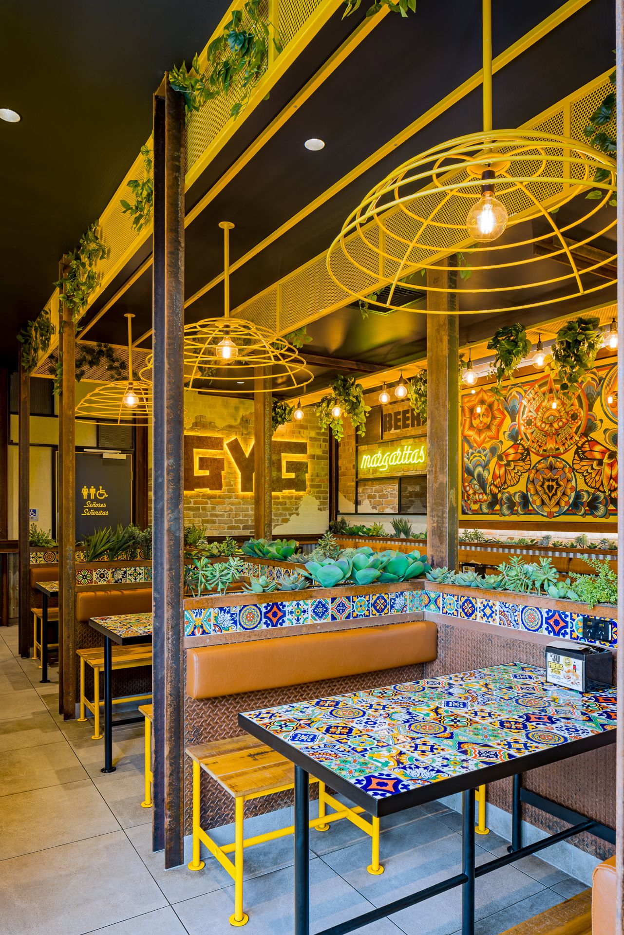 Colourful Mexican restaurant interior with tiled tables, tan leather booths, yellow stools, hanging plants and wire cage pendant lights.