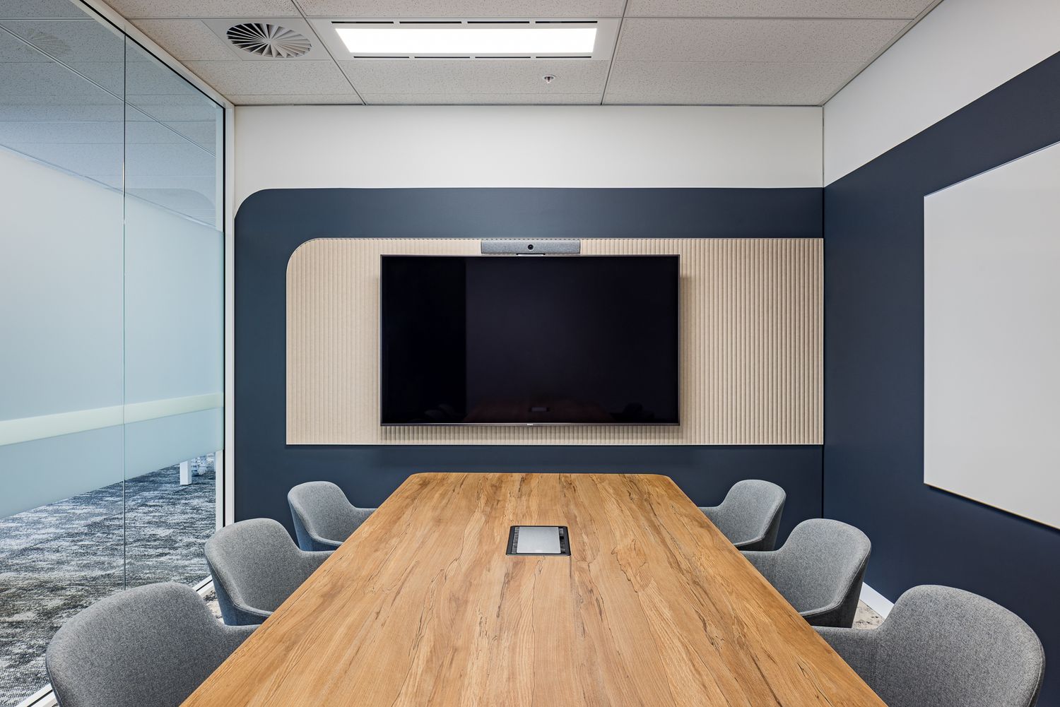 Modern meeting room with a long timber table, grey chairs, wall-mounted display screen, navy accent panelling and a whiteboard.