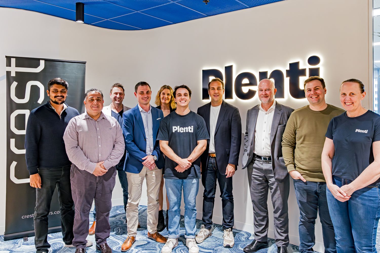 Eleven smiling colleagues pose together in front of an illuminated Plenti sign on a wall in a modern office.