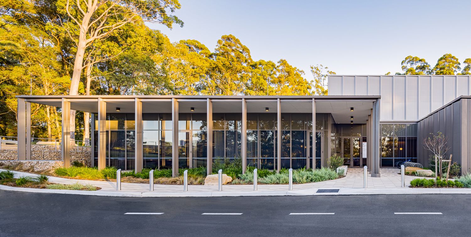 Glass-walled building entrance with slender steel columns, native garden beds and a footpath surrounded by tall eucalyptus trees.