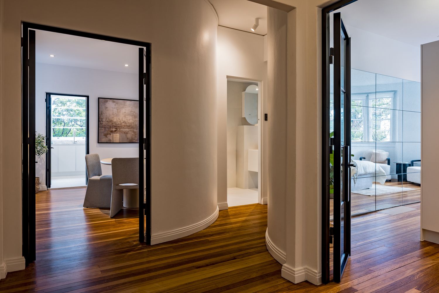 Bright hallway with curved walls and polished timber floors leading to a sitting room, a dining area and a mirrored bedroom.