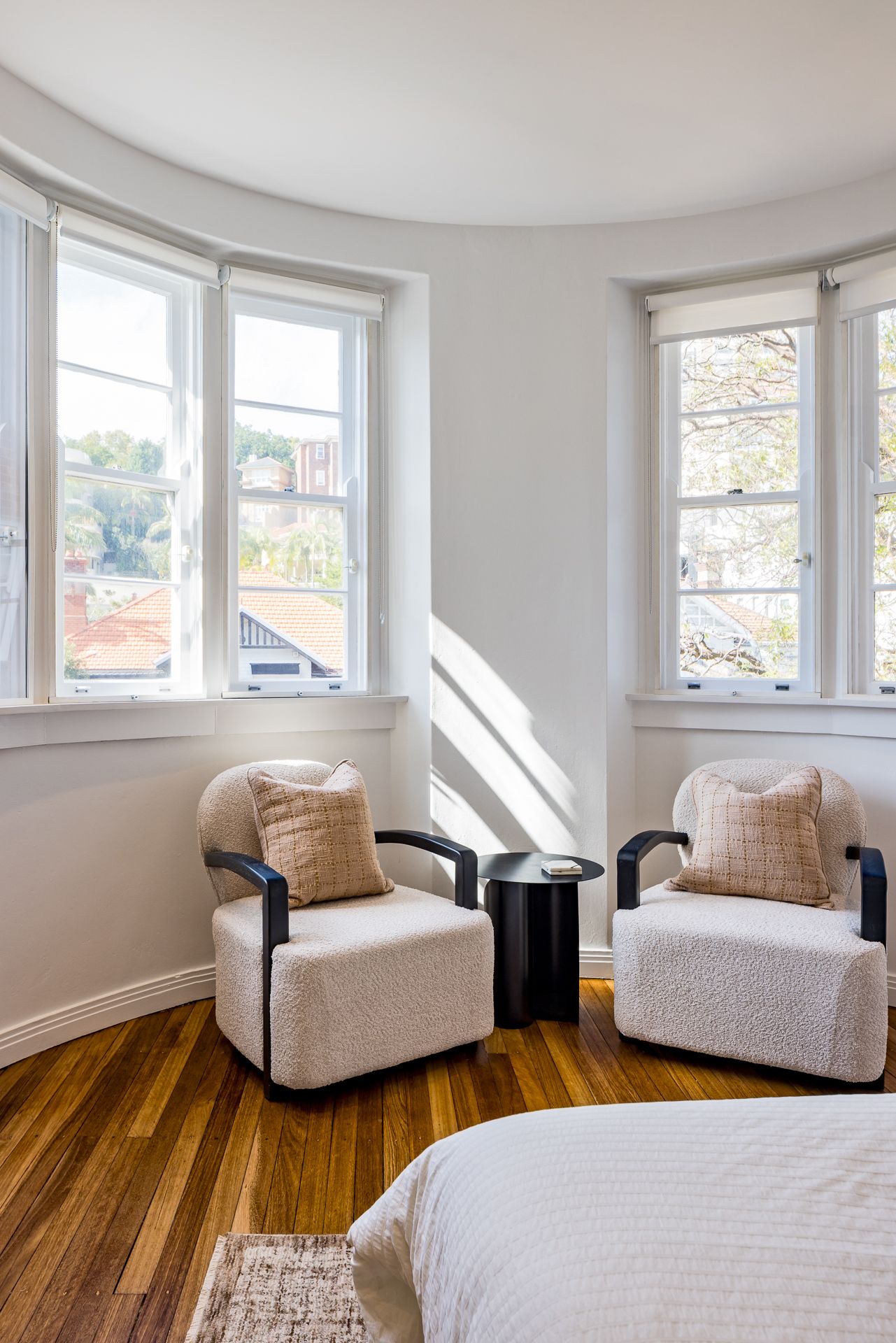 Two cream boucle armchairs with a black side table in a sunny curved window nook above timber floors.