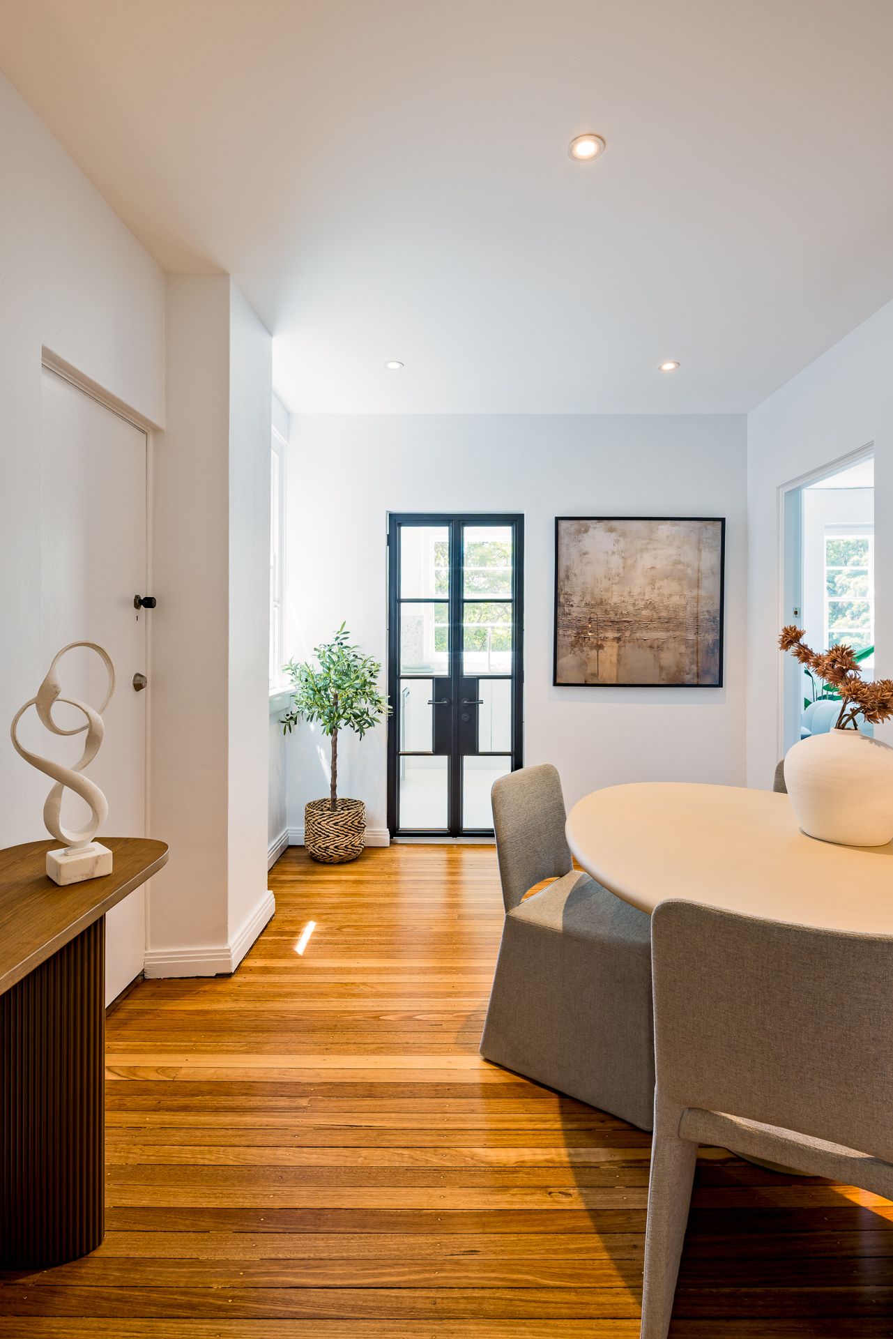 Bright open-plan dining space with round white table, grey chairs, sculptural console, abstract art and timber floors.