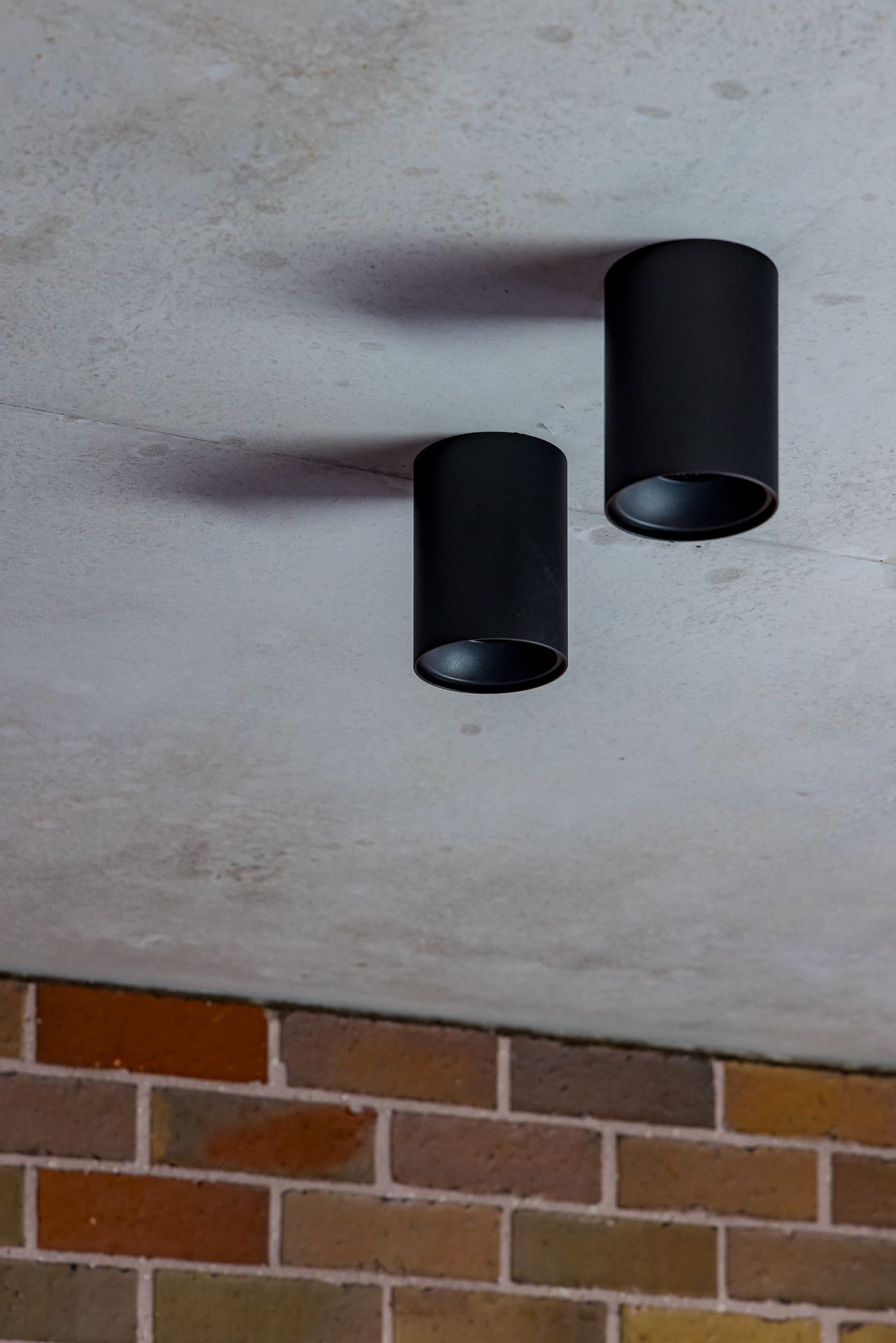 Two black cylindrical surface-mounted downlights on a raw concrete ceiling above a textured brick wall.