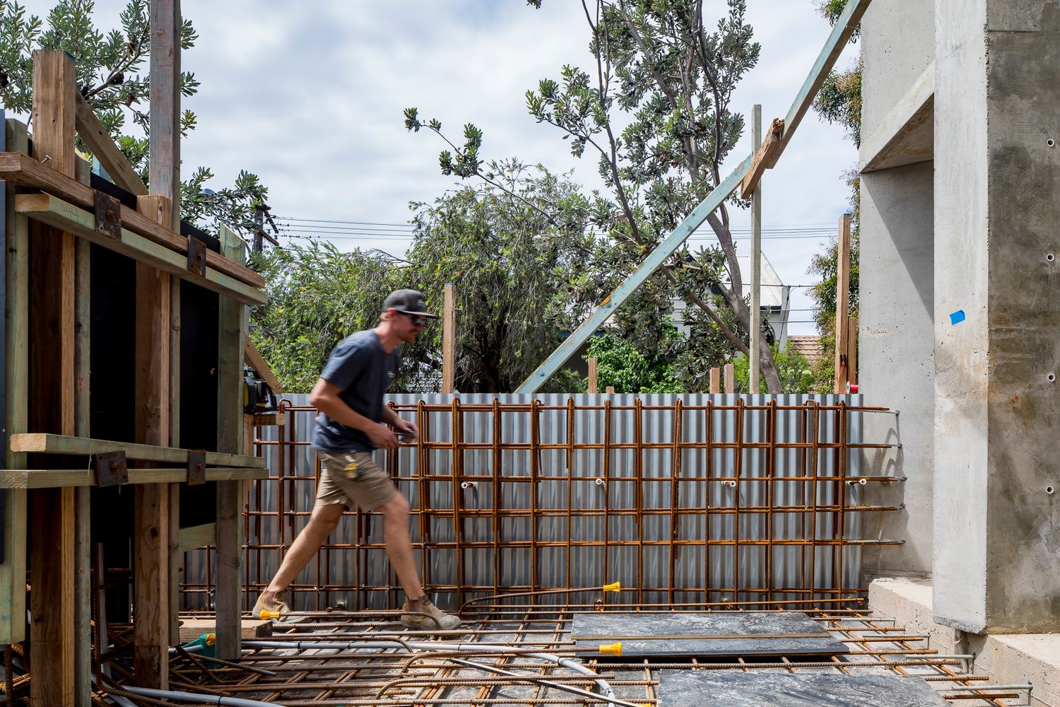 Construction worker walking across a building site with timber formwork, steel reinforcement bars and concrete walls beneath trees.