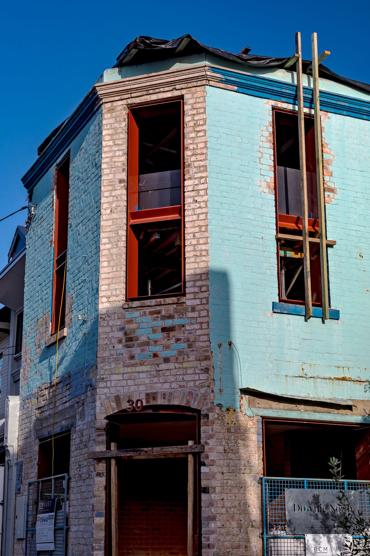 Corner brick building under renovation with blue painted walls, red window frames, and scaffolding poles against a clear sky.