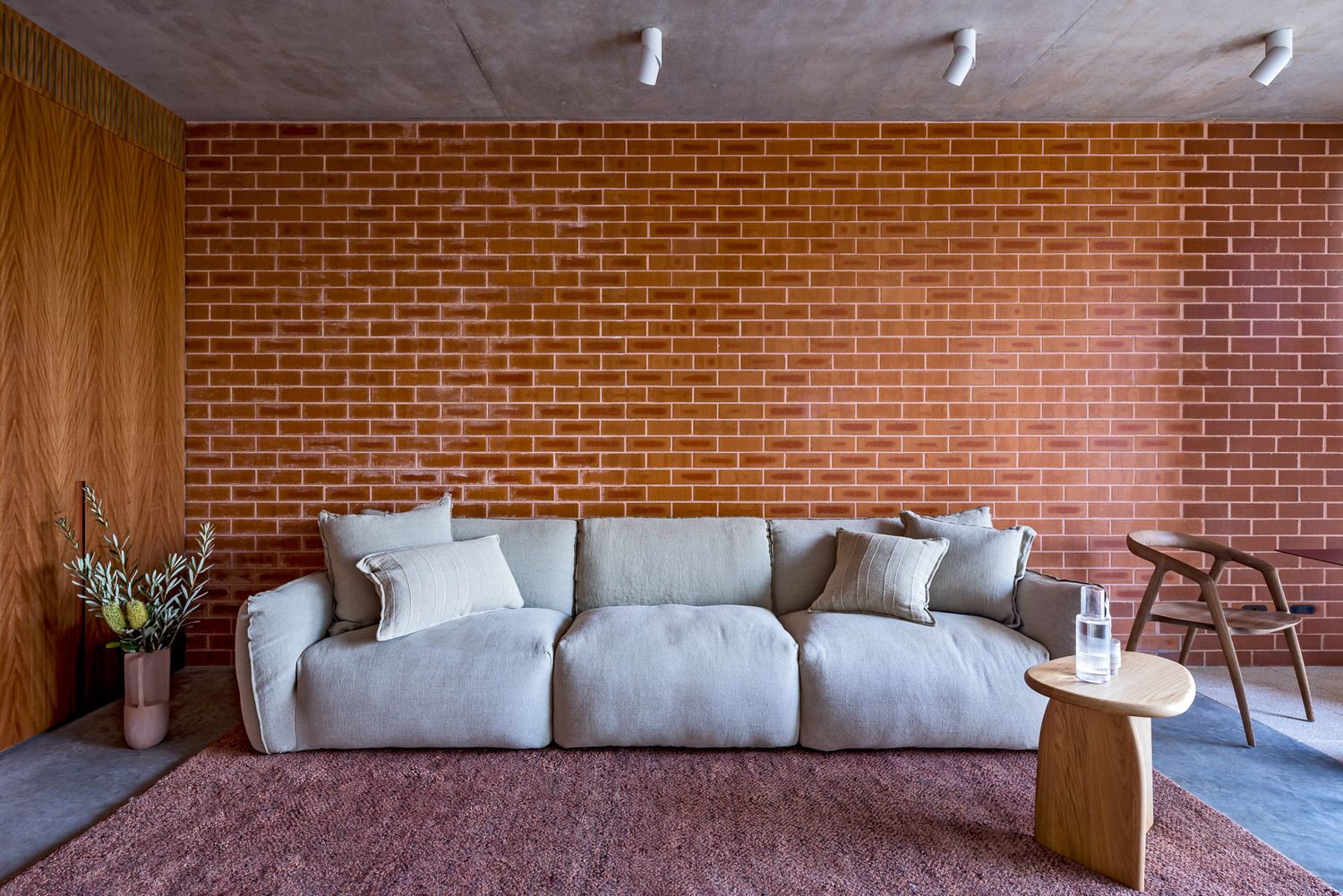 Modern living room featuring a pale gray sectional sofa, red brick feature wall, timber panelling, a wooden chair and a dusty rose rug.