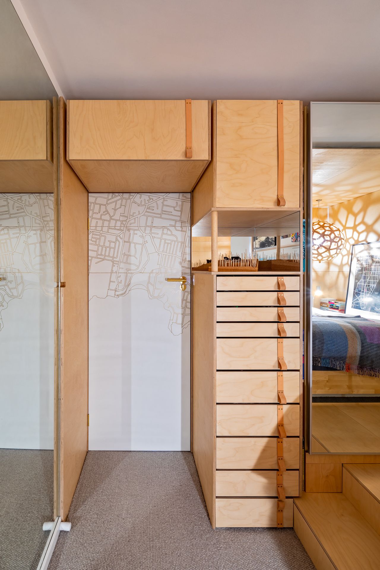 Plywood storage unit with leather-pull drawers beside a white door printed with a map pattern and a bedroom step.
