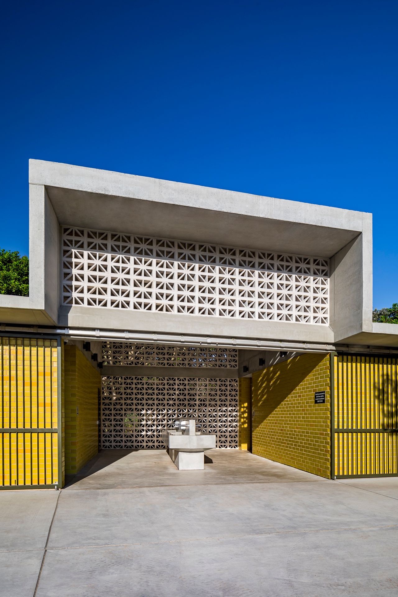 Modern concrete building entrance with a geometric breeze block facade, yellow slatted gates and a stone fountain under a clear blue sky.